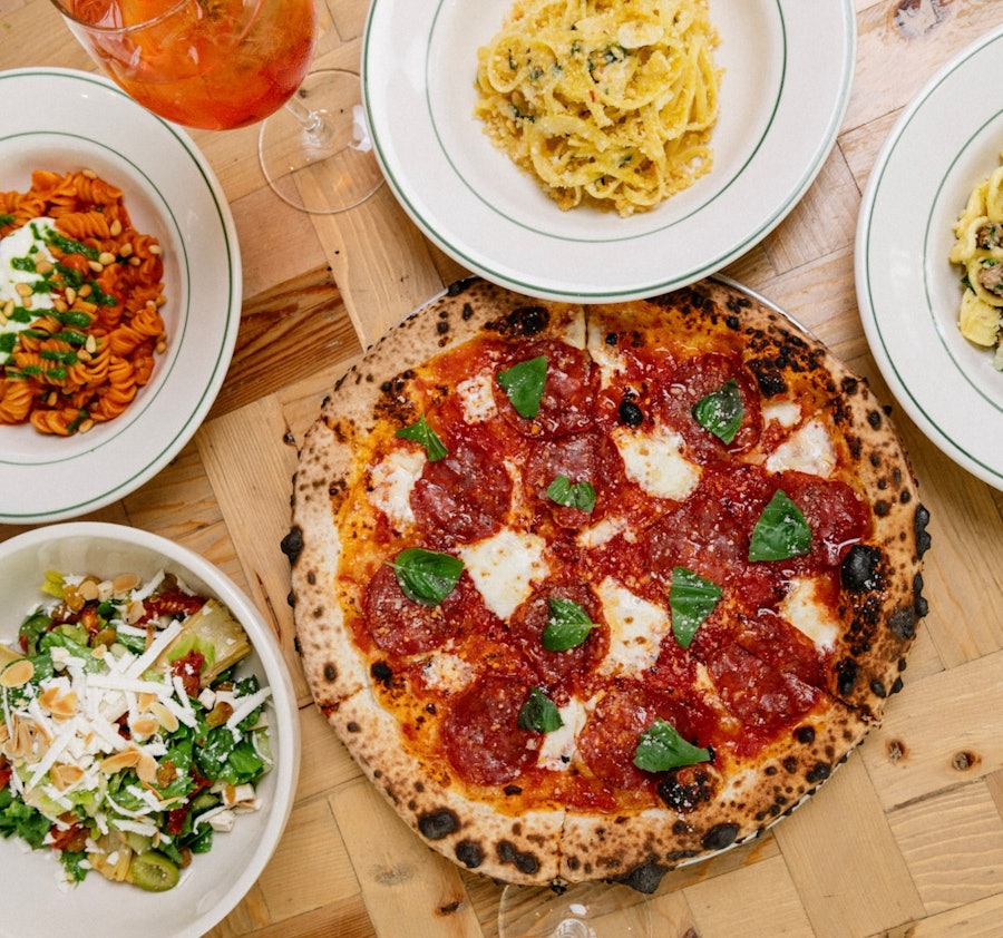 A spread of Rosemary's East dishes including pasta, salad, spritz cocktails, and a pizza topped with basil and soppressata.