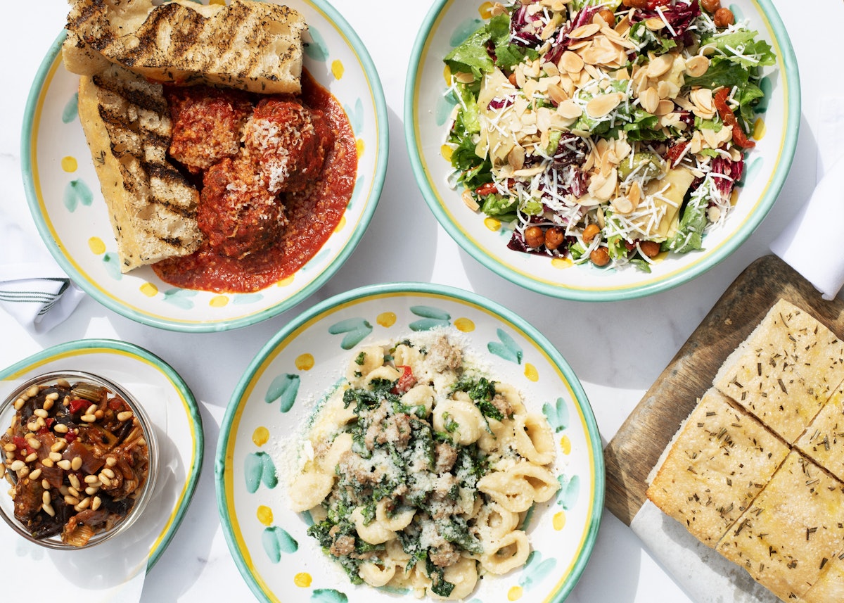 Overhead image of signature dishes from Rosemary's: pastas, salads, meatballs, and focaccia, served in painted porcelain plateware.